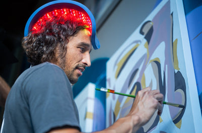 Male painter wearing a Celluma RESTORE red light therapy device on his head to stimulate hair growth while painting on a canvass