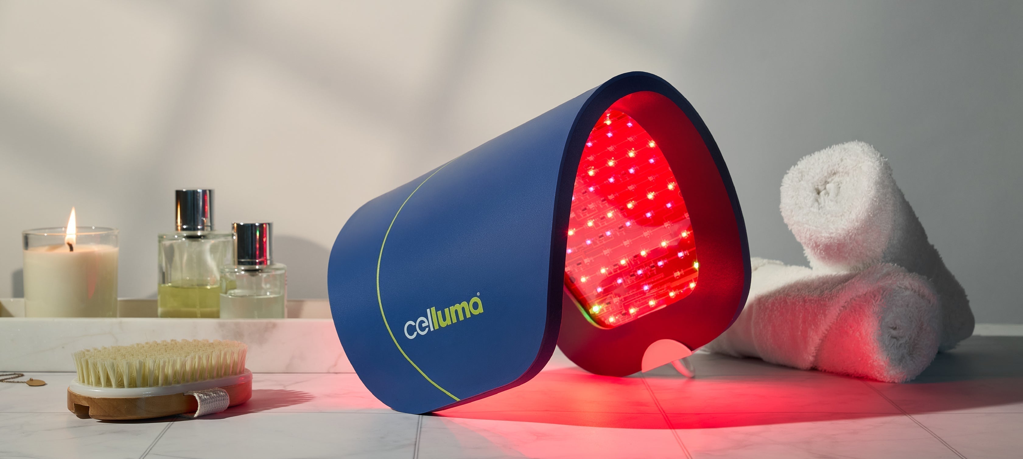 A Celluma panel in a curved shape sitting on a table with two small, rolled towels, a body brush, a lit candle, and two bottles of body oil
