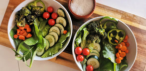 Two bowls filled with sliced cucumbers, carrots, peppers, tomatoes, broccoli, and lettuce with a small bowl of salt on top of a cutting board