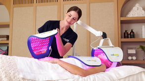 Esthetician placing a panel onto a clients hands while they lie on a massage table with additional panels over their neck/chest and face from the Celluma ELITE device.