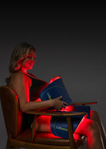 Woman sitting in chair receiving a LED red light therapy body contouring treatment on her thighs and abdomen using a Celluma CONTOUR PLUS multi-panel LED device.