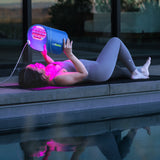 Woman lying by the pool using a Celluma PRO light therapy device at dusk