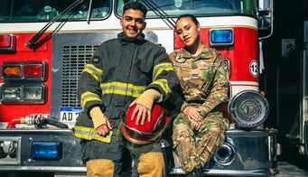 Two firefighters, one in a fire helmet and the other in camouflage, standing in front of a fire truck.